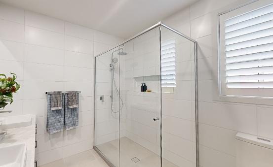 A modern, white-tiled bathroom with a glass-enclosed shower, two blue towels hanging on the wall, and a window with white shutters