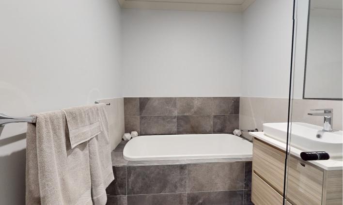 A modern bathroom with a built-in bathtub, gray tiled walls around the tub, light-colored towels on a towel rack, and a wooden vanity with a white sink and chrome faucet