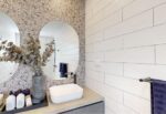 A modern bathroom a terrazzo accent wall with two round mirrors, a minimalist white sink, and a countertop decorated with a vase of dried branches and toiletries