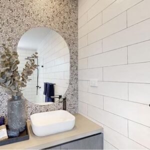 A modern bathroom a terrazzo accent wall with two round mirrors, a minimalist white sink, and a countertop decorated with a vase of dried branches and toiletries