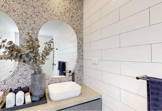 A modern bathroom a terrazzo accent wall with two round mirrors, a minimalist white sink, and a countertop decorated with a vase of dried branches and toiletries