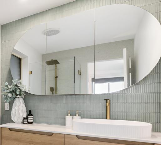 Modern bathroom features a large oval mirror, a sleek white sink with a gold faucet, and decorative accessories on a wood vanity against a green-tiled wall